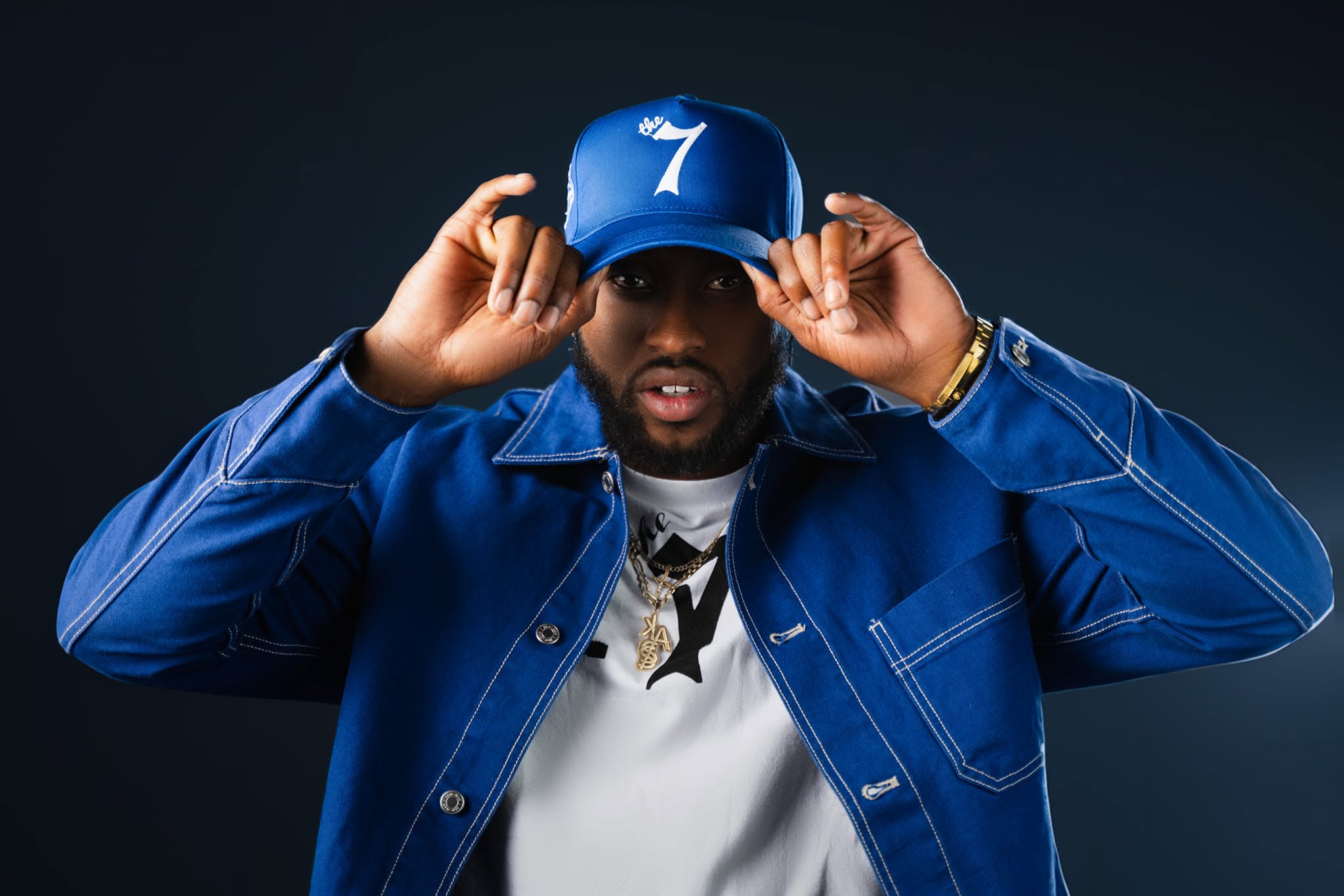 JayDubb Tha Ruler adjusts a blue The 7 cap in a studio portrait